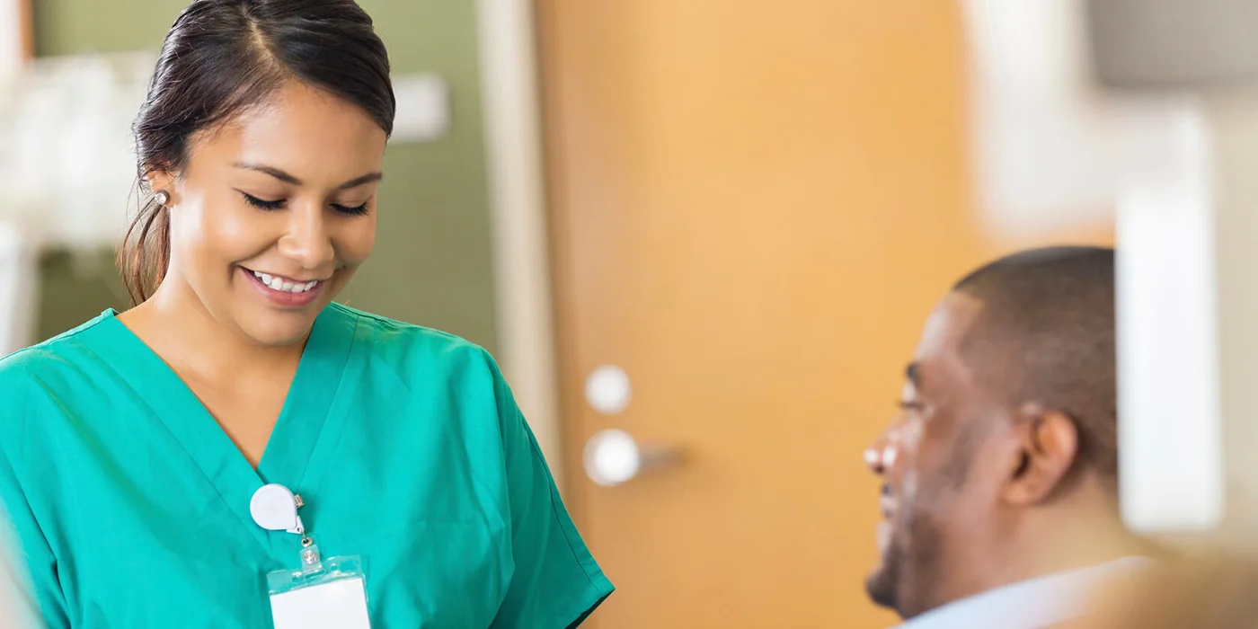 A nurse in scrubs smiling as she speaks with a resident at the bedside