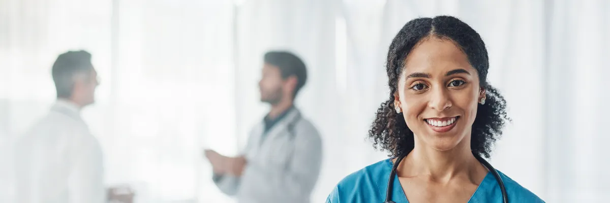 A smiling nurse in scrubs, two clinicians talking in the background