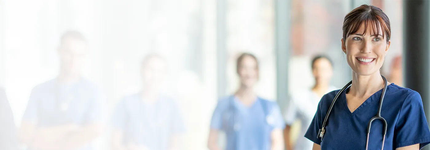 A nurse in navy scrubs with a stethoscope, colleagues out of focus behind her
